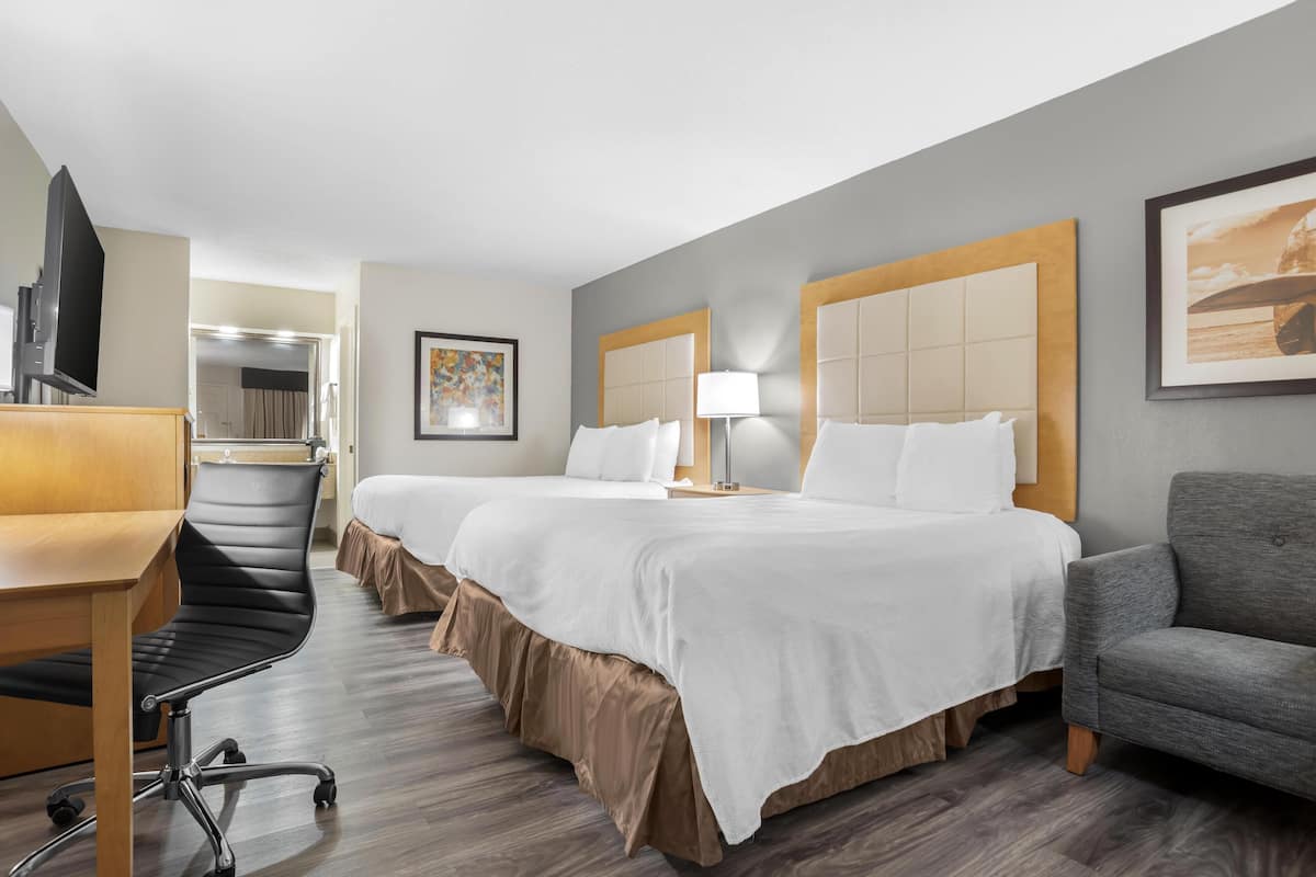 A hotel room with two beds, a desk, a chair, a TV, and a lamp.