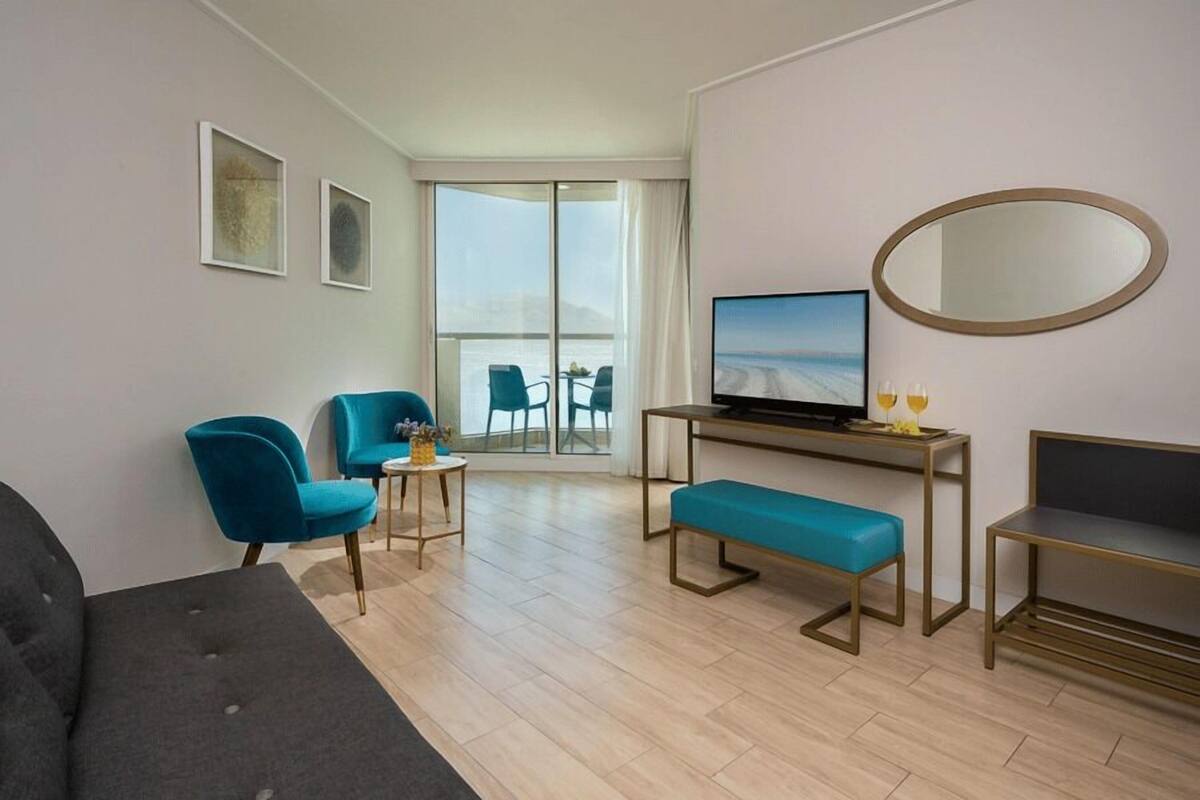 Una habitación de hotel moderna con un sofá, dos sillones azules, una mesita con un televisor, un banco y un amplio ventanal con vistas.