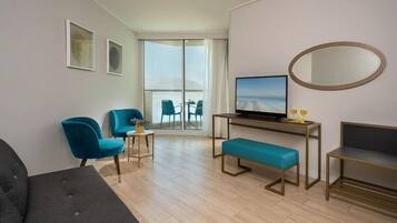 A modern hotel room with a sofa, two blue armchairs, a small table with a TV, a bench, and a large window with a view.