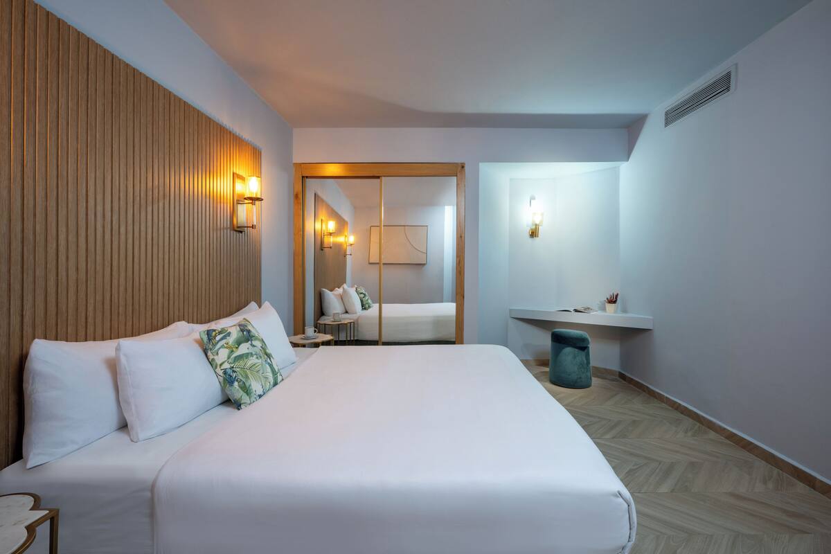 Habitación de hotel con una cama grande, cabecera de madera, mesita de noche, tocador, espejo, luz adosada a la pared, un taburete pequeño y un lavabo con encimera.