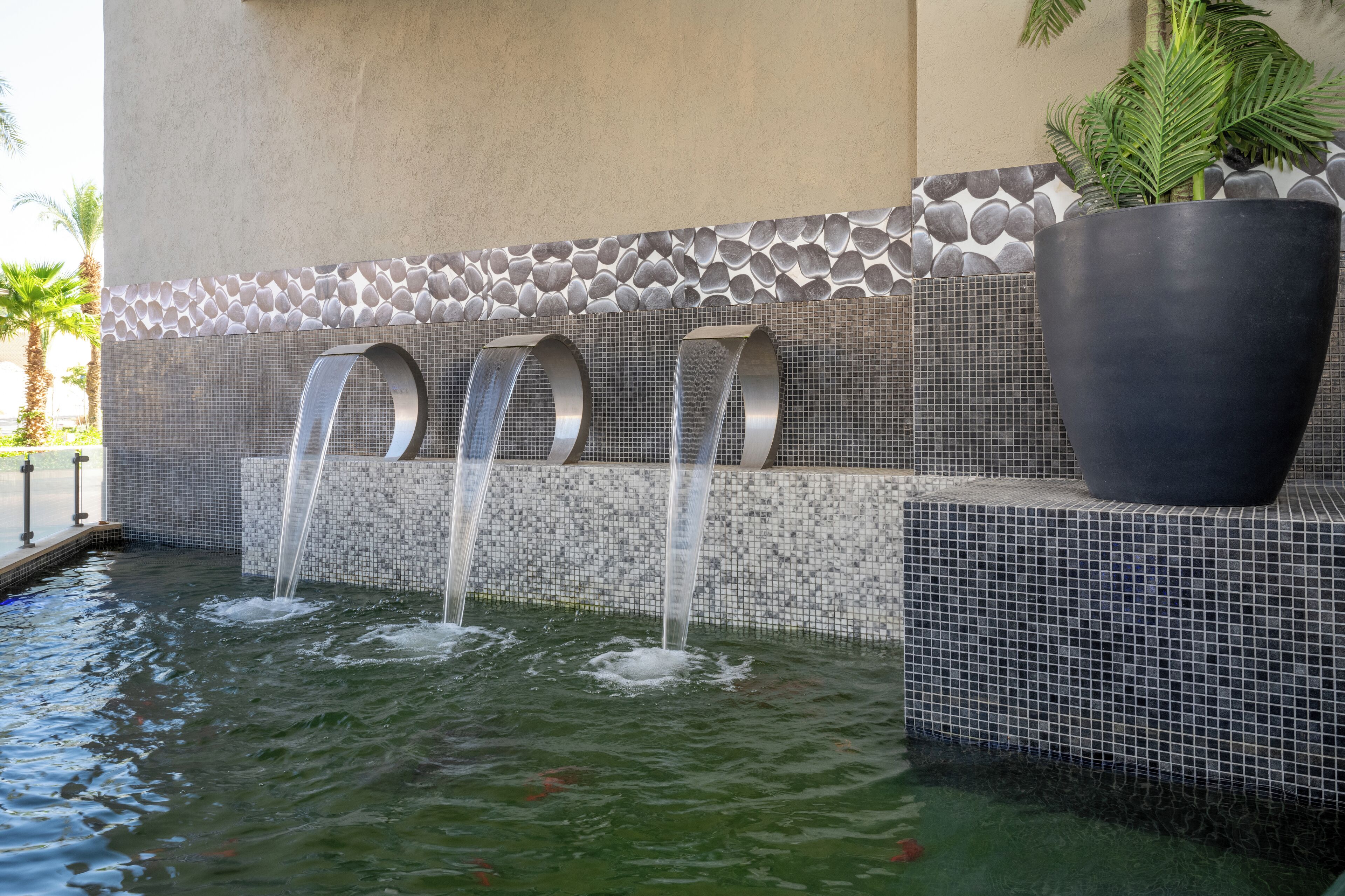 A water feature with three curved spouts, a large planter, and a tiled wall with a pebble pattern.