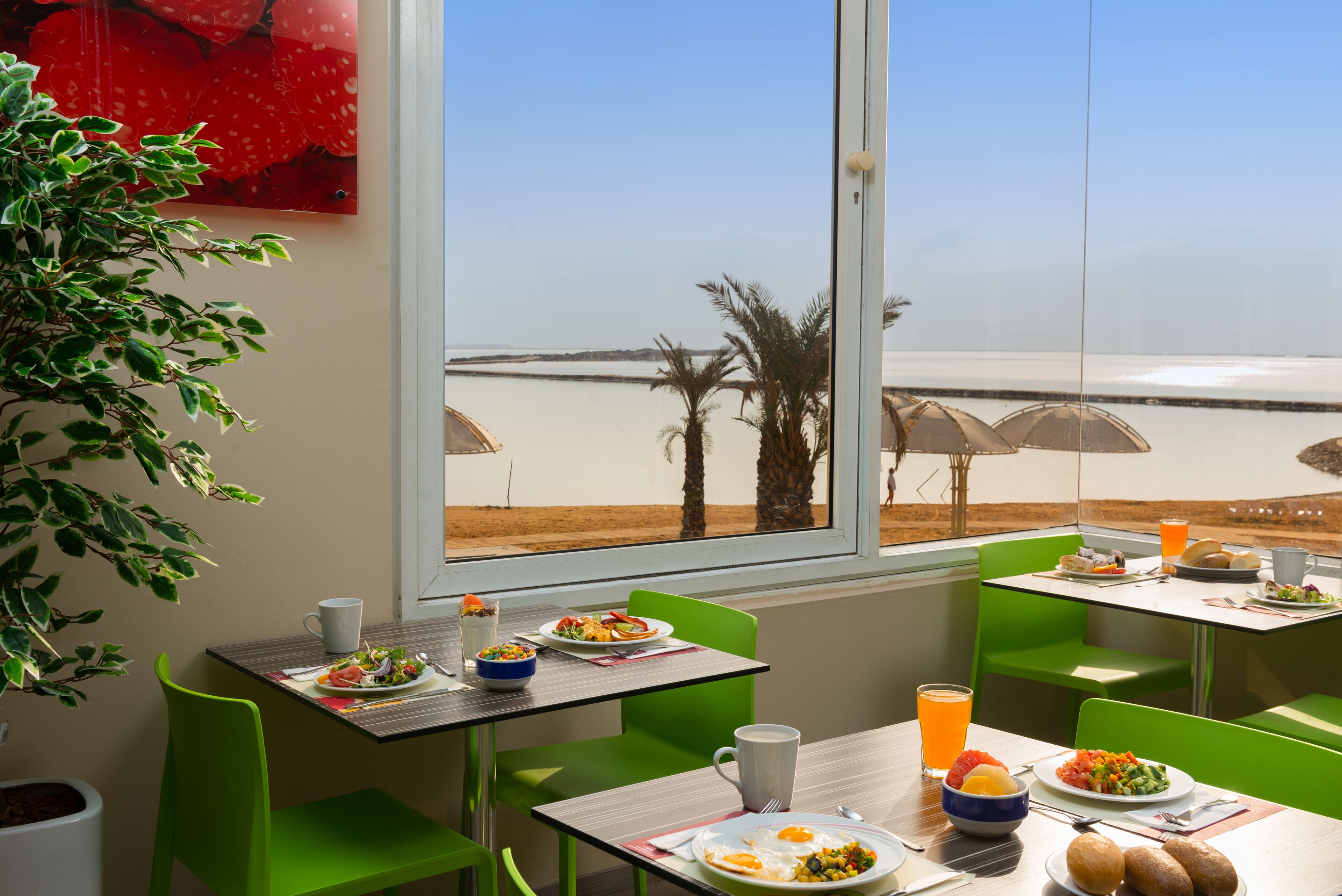 Comedor con vista a la playa, sillas y mesas verdes dispuestas con platos de desayuno y bebidas.