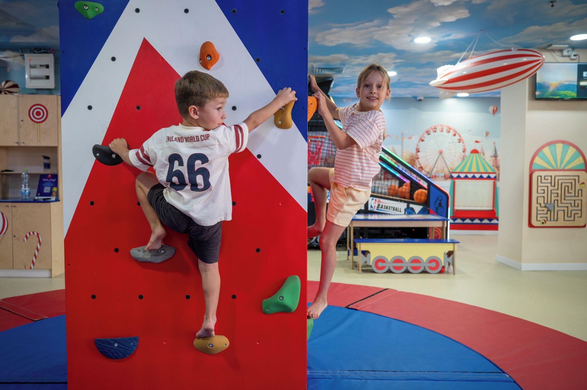 A climbing wall with a red and white design, a basketball court, and a Ferris wheel in the background.