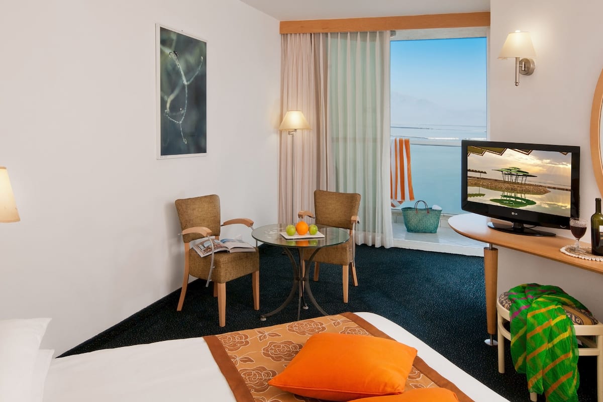 Habitación de hotel con cama, escritorio con televisor, dos sillas, una mesita con frutas y ventana con cortinas.