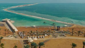 Luftaufnahme eines Strandresorts mit einer langen Sandküste, klarem türkisblauem Wasser und vereinzelten Palmen. Sonnenschirme und Liegestühle sind für die Gäste bereitgestellt.