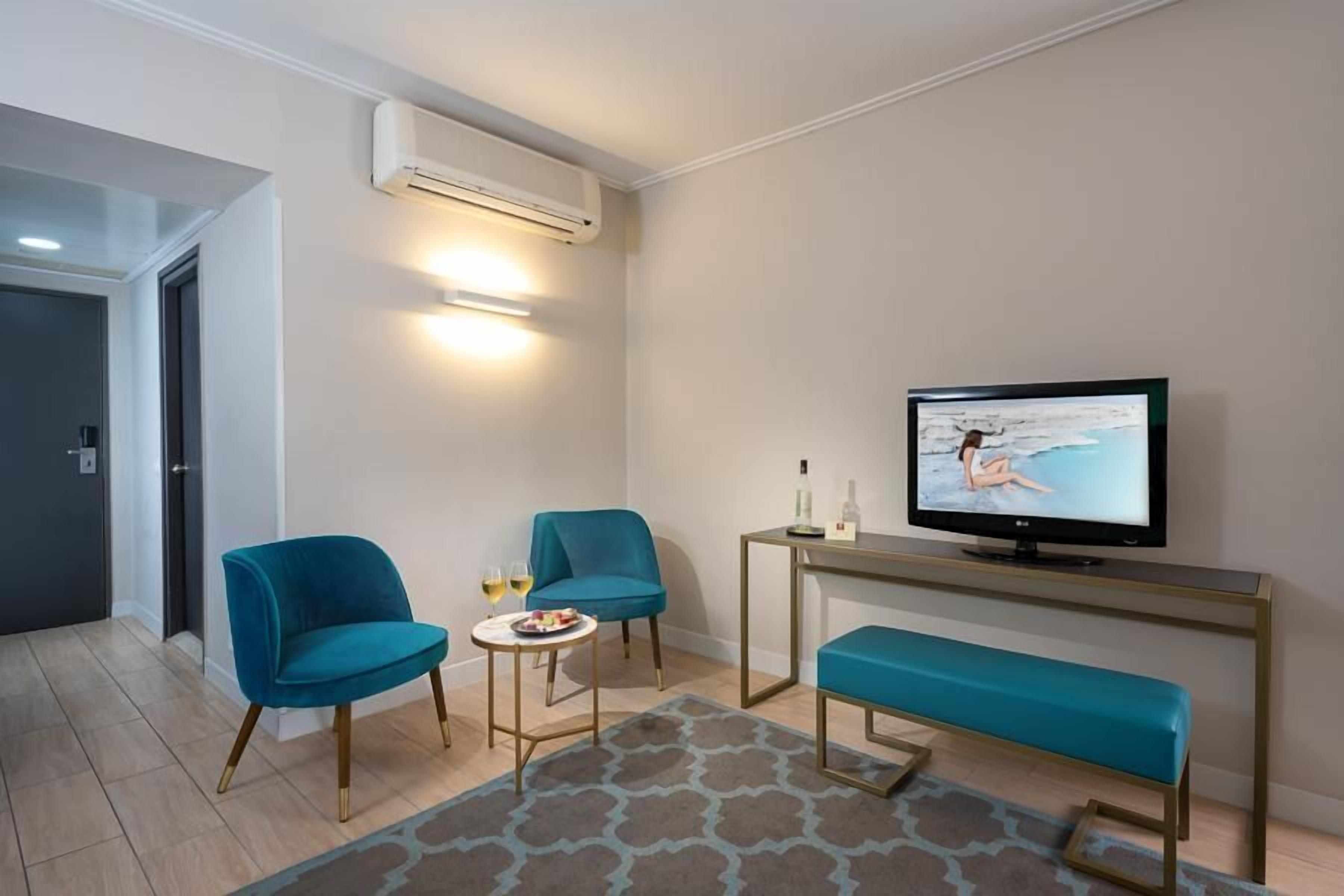 A modern living room with a TV, two blue chairs, a small table with drinks, and a bench.