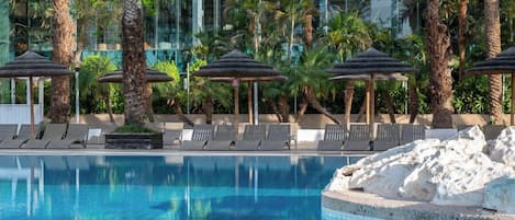 A pool with lounge chairs and umbrellas, surrounded by palm trees and a modern building.