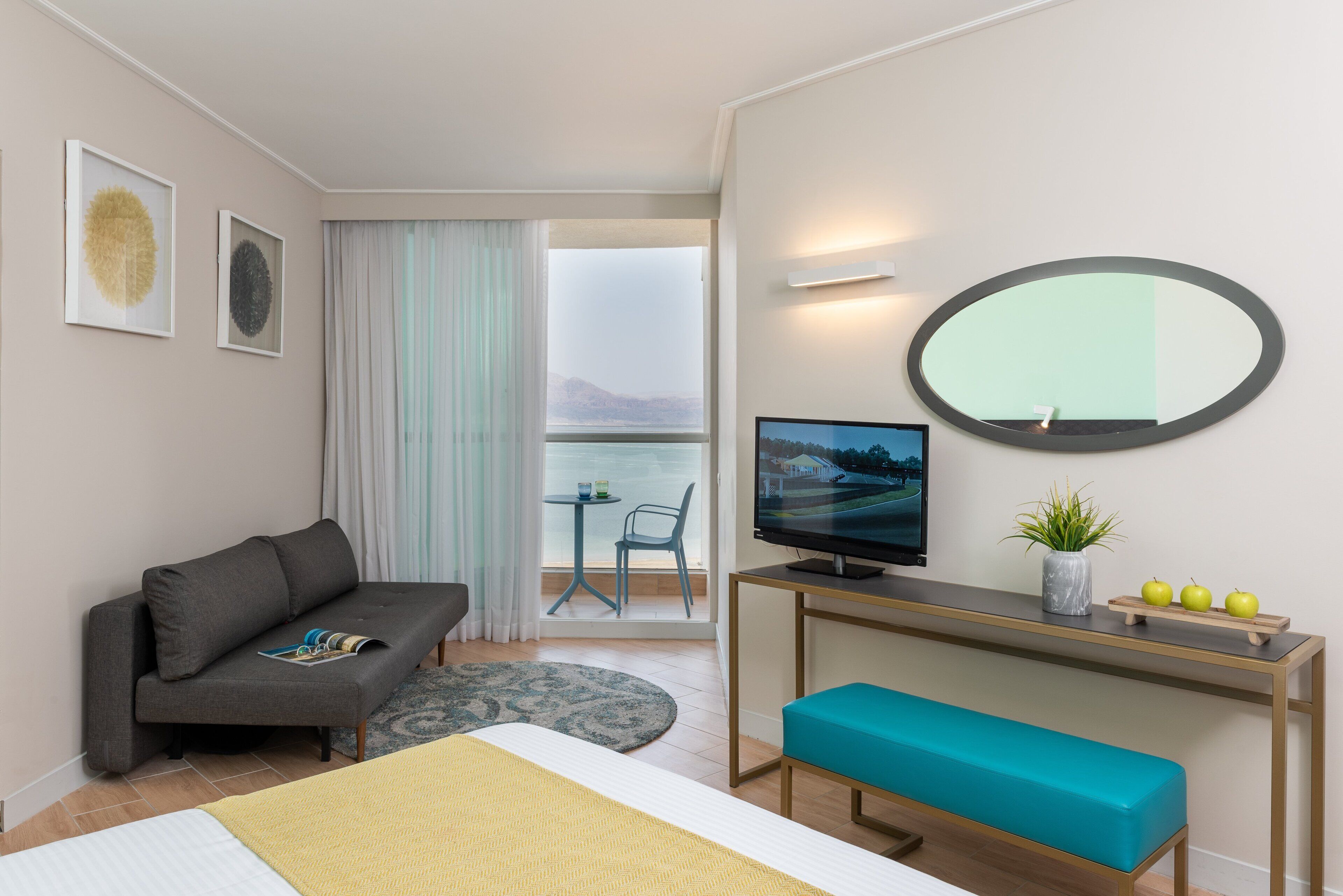 A modern hotel room with a sofa, a bed, a desk with a TV, a bench, a small table with apples and a plant, and a balcony with a view.