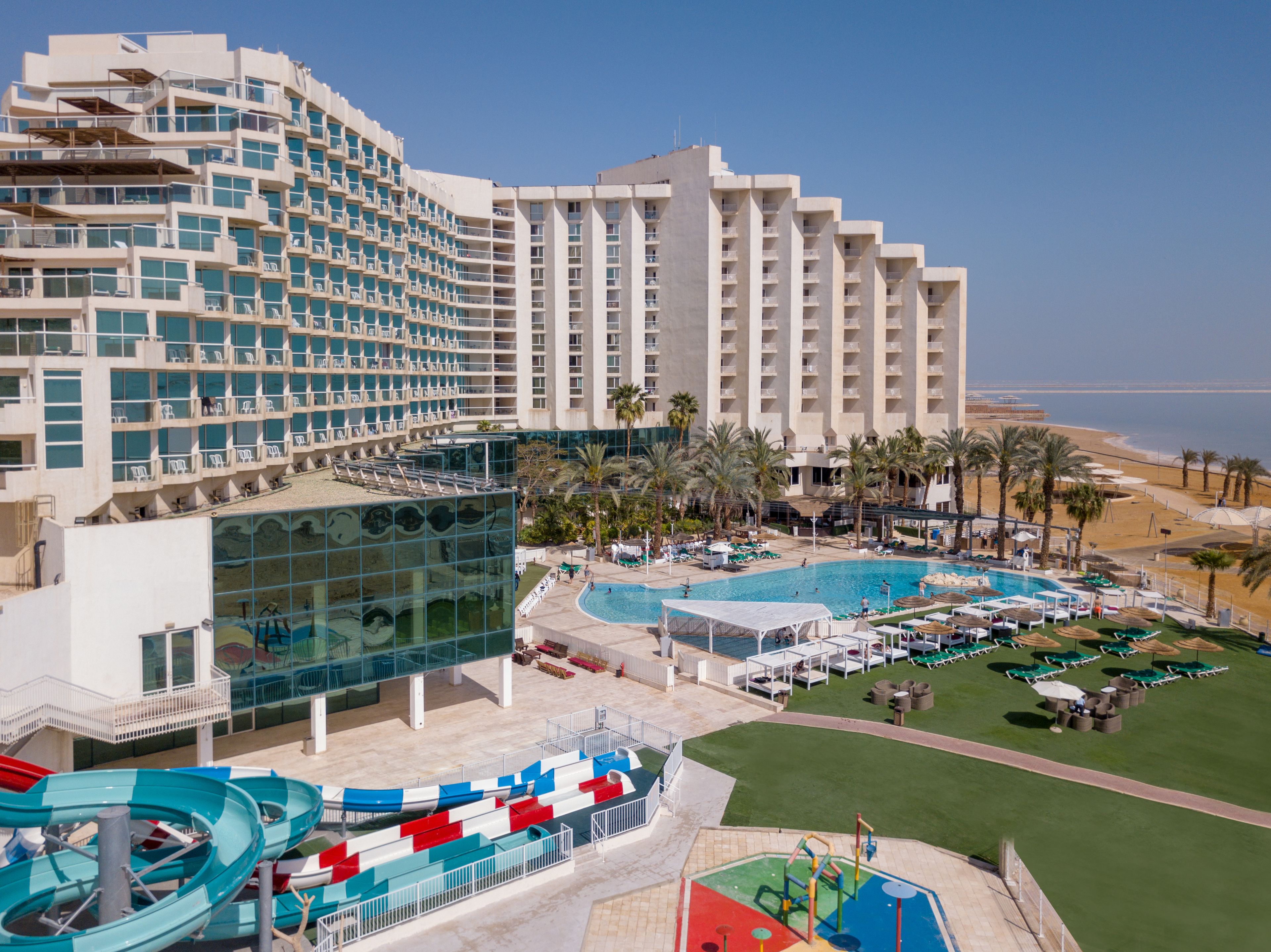 A hotel with multiple floors, a pool area with lounge chairs, a water slide, and palm trees.