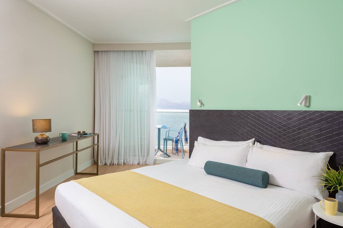 Habitación de hotel con cama, mesita de noche con lámpara, vista al mar y balcón con silla y mesa.