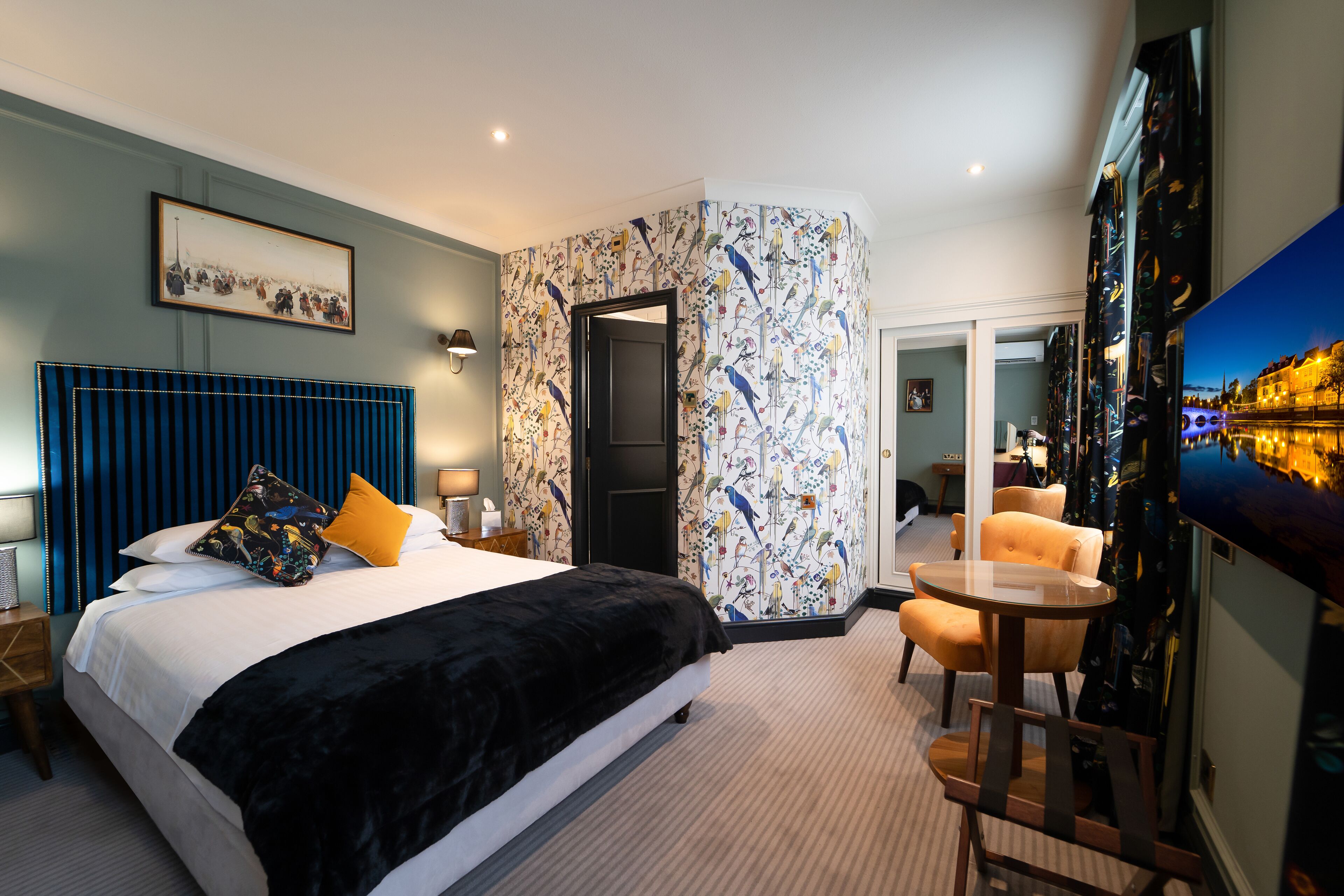 Habitación de hotel con una cama grande, un rincón de estar con una mesa y sillas, un televisor instalado en la pared y una puerta que da a otra habitación.
