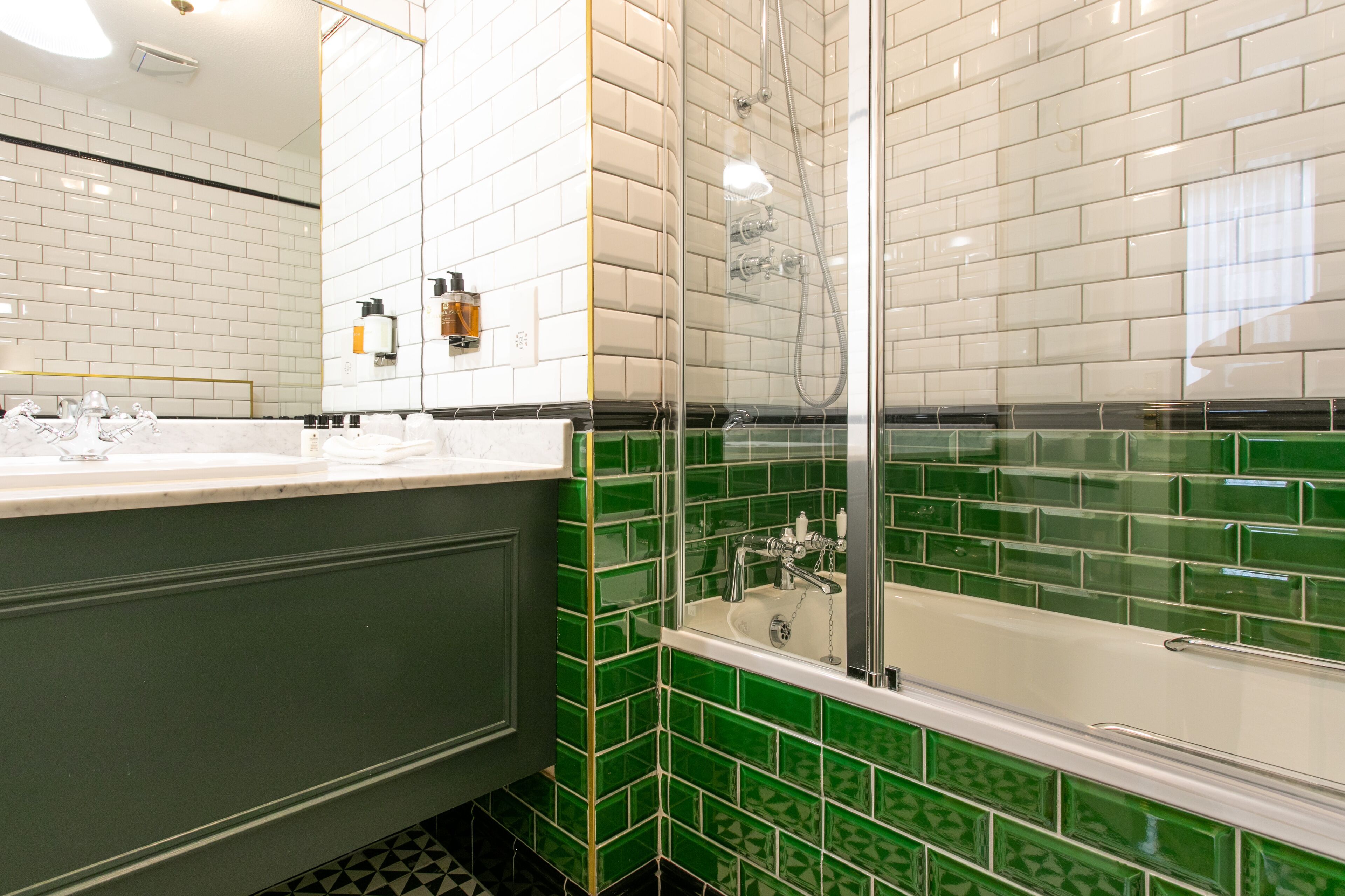 Un baño con pared de azulejos blancos, zona de ducha con azulejos verdes y un lavamanos blanco con espejo.