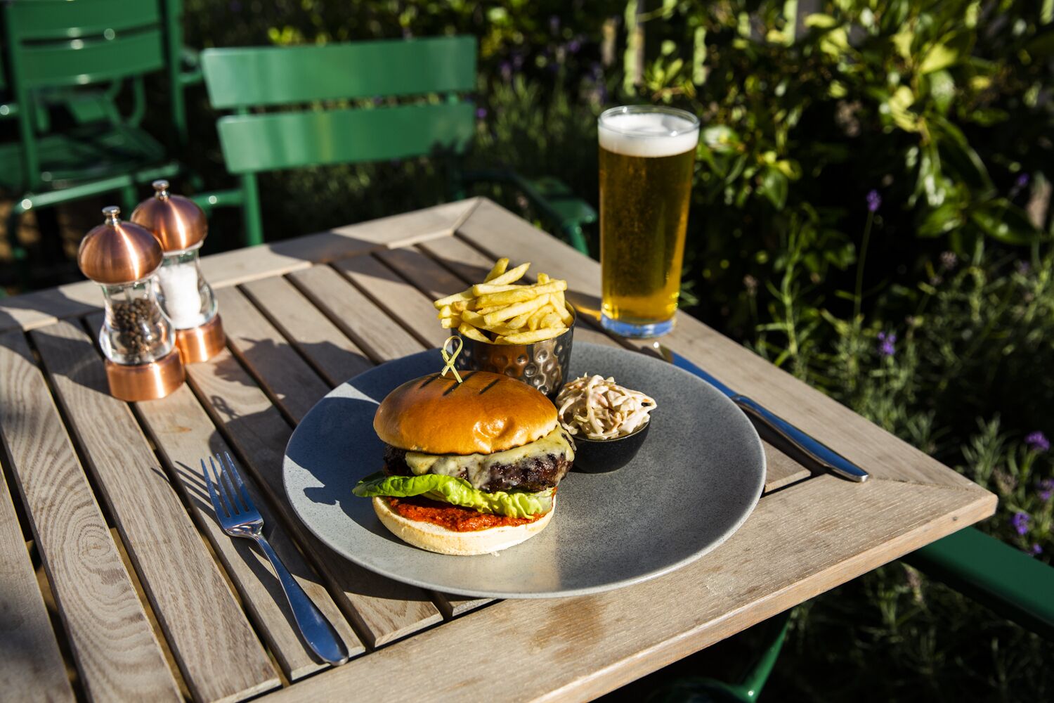 En måltid bestående av en hamburgare, pommes frites, coleslaw och ett glas öl, serverad på ett träbord utomhus.
