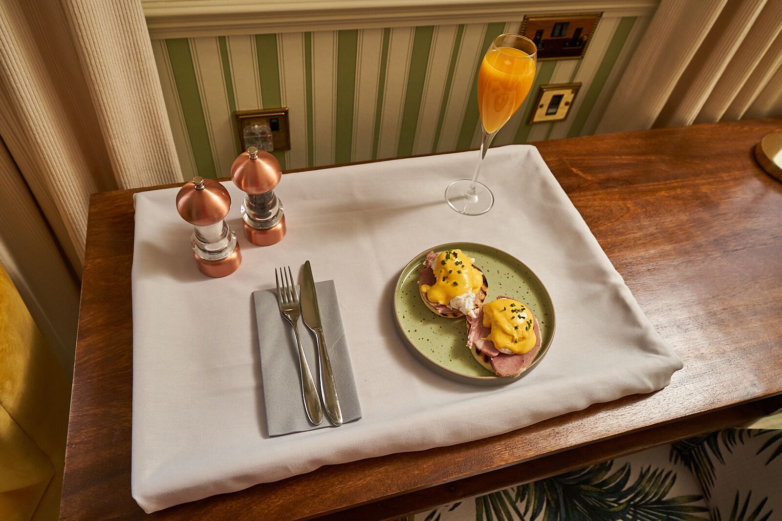 A breakfast setup with two eggs benedict on a green plate, a glass of orange juice, and a set of silverware on a white tablecloth.