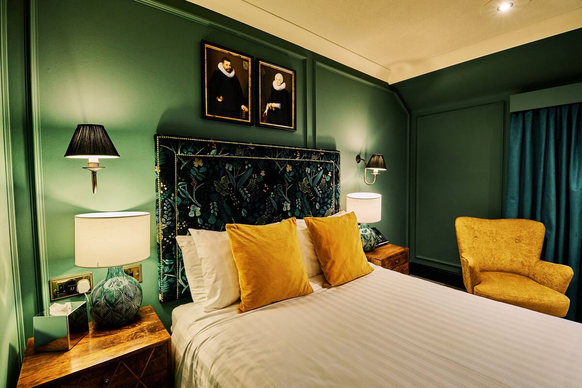 Habitación de hotel ordenada con una cama grande, dos lámparas de noche, un cabecero decorativo y un sillón amarillo.