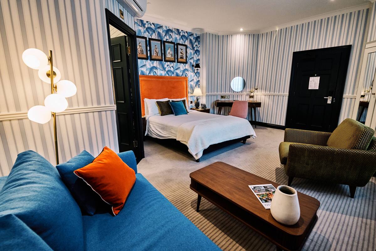 Una habitación de hotel con una cama, un sofá azul, una mesa de centro de madera, un sillón verde y una pared a rayas con motivos tropicales.
