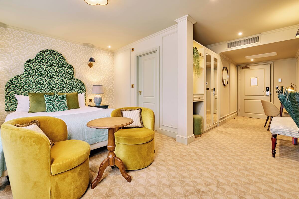 Habitación de hotel con cama, dos sillones amarillos, una mesita redonda, un espejo y una puerta que da a otra habitación.