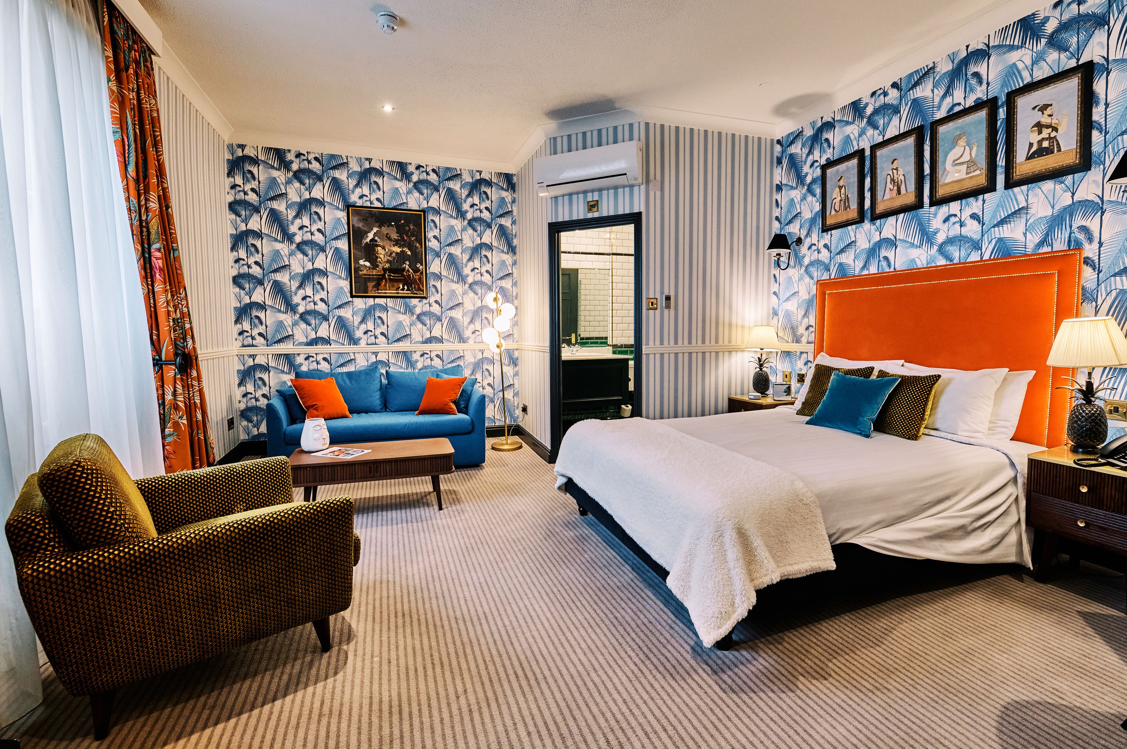 Une chambre d’hôtel avec un grand lit, un canapé, un fauteuil et une petite table. On y trouve une porte donnant sur une salle de bain et un mur orné de tableaux.