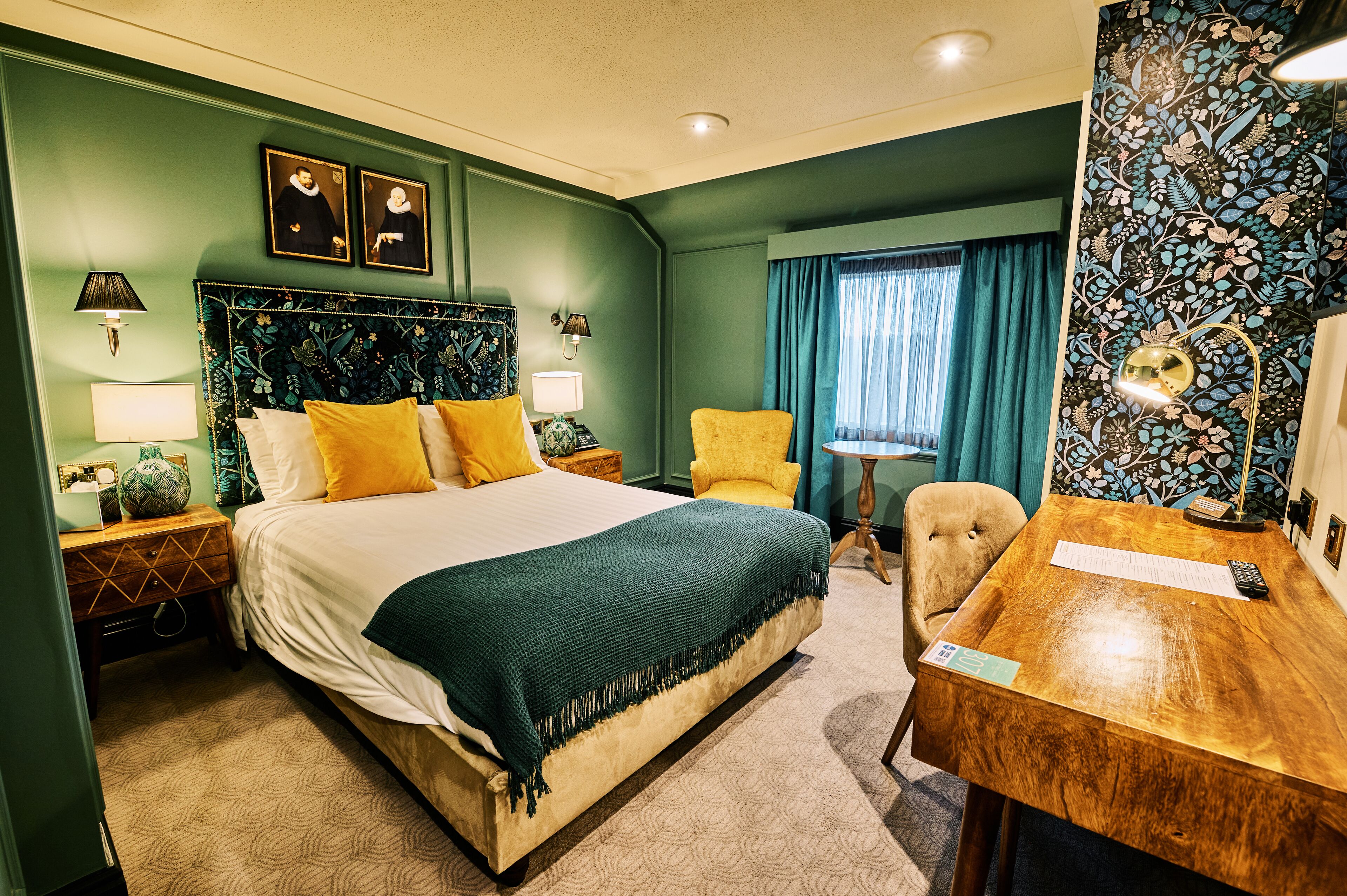 Quarto de hotel com uma cama grande, uma escrivaninha de madeira, uma cadeira e uma luminária de mesa.