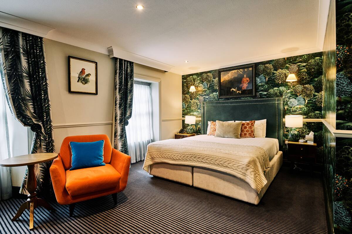 Habitación de hotel con una cama grande, una mesita de noche, una silla, una pequeña mesa y un mural temático de naturaleza en la pared.