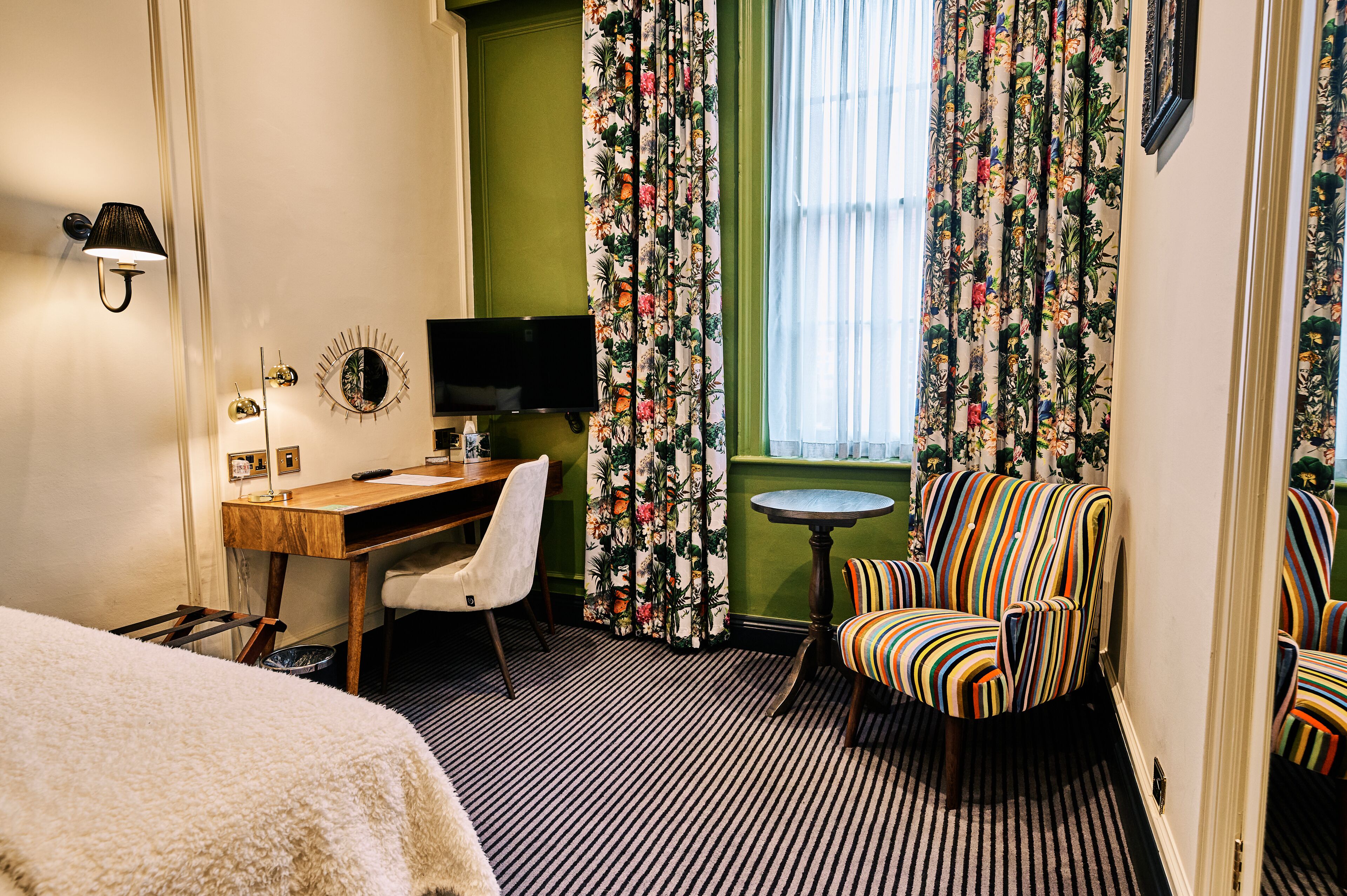 A hotel room with a bed, a desk with a chair, a television, a small round table, and two striped armchairs.