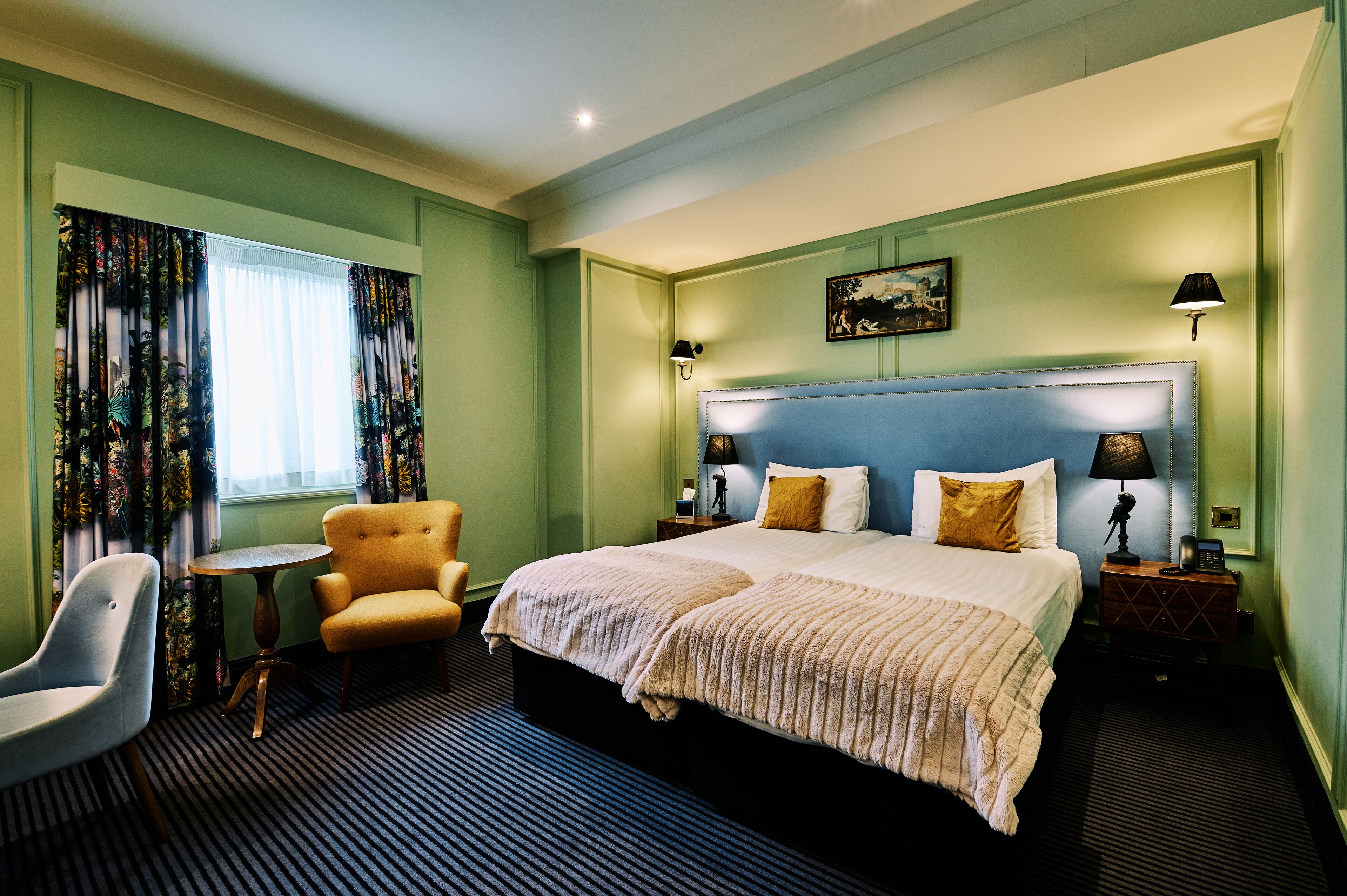 A hotel room with a large bed, two bedside tables with lamps, a chair, a small round table, and curtains with a tropical print.