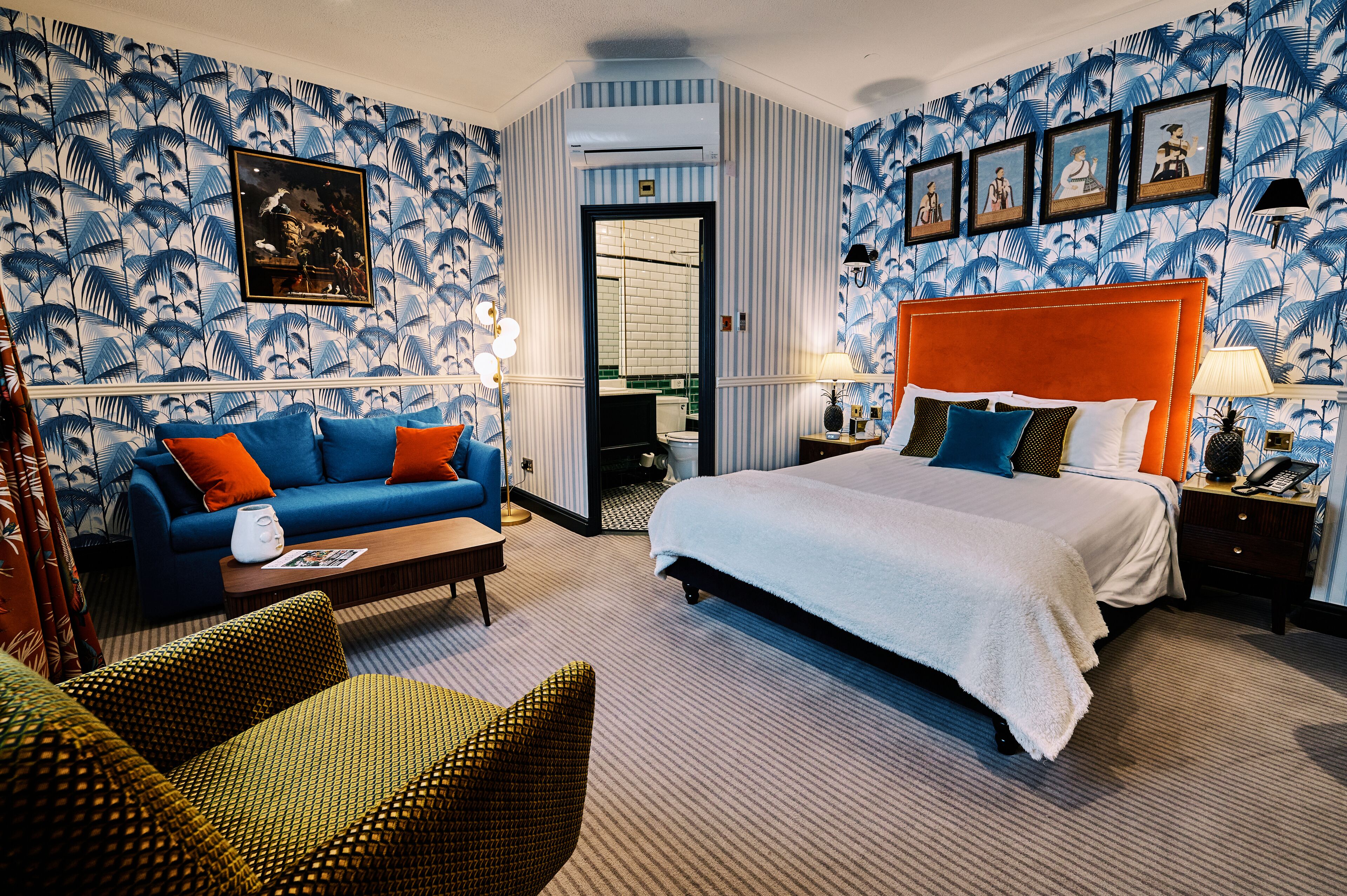 A hotel room with a large bed, a sofa, a coffee table, and a chair. There is a bathroom visible through an open door.