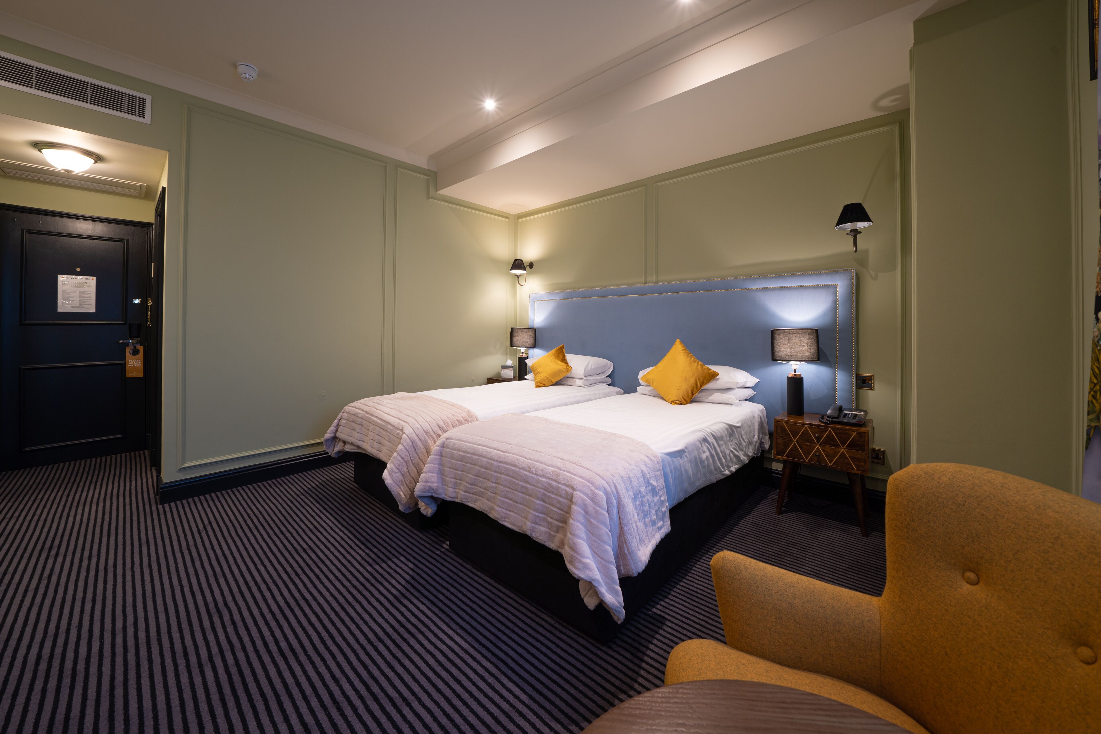 A hotel room with two beds, a nightstand, a chair, and a lamp.