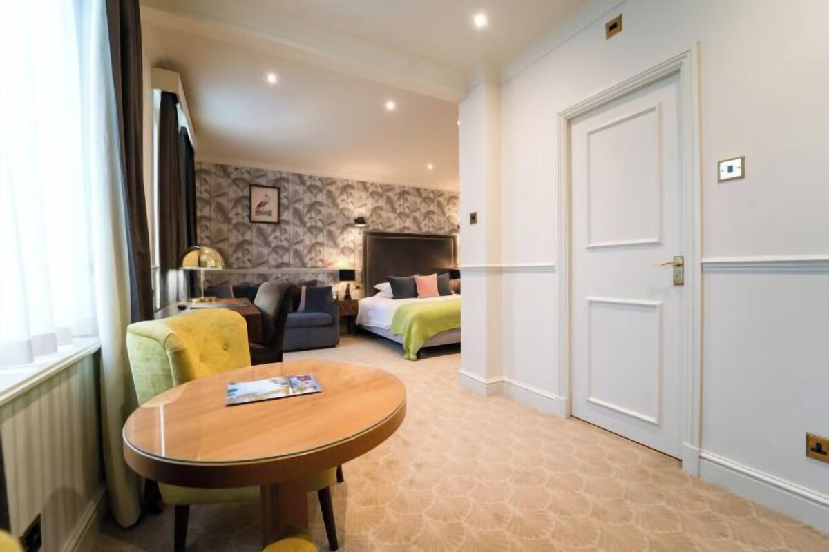 A hotel room with a round wooden table, a sofa, a bed with a green blanket, and a patterned wallpaper.