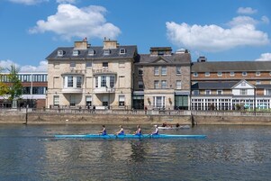 A row of traditional buildings along a river, with a rowboat with rowers in the foreground.