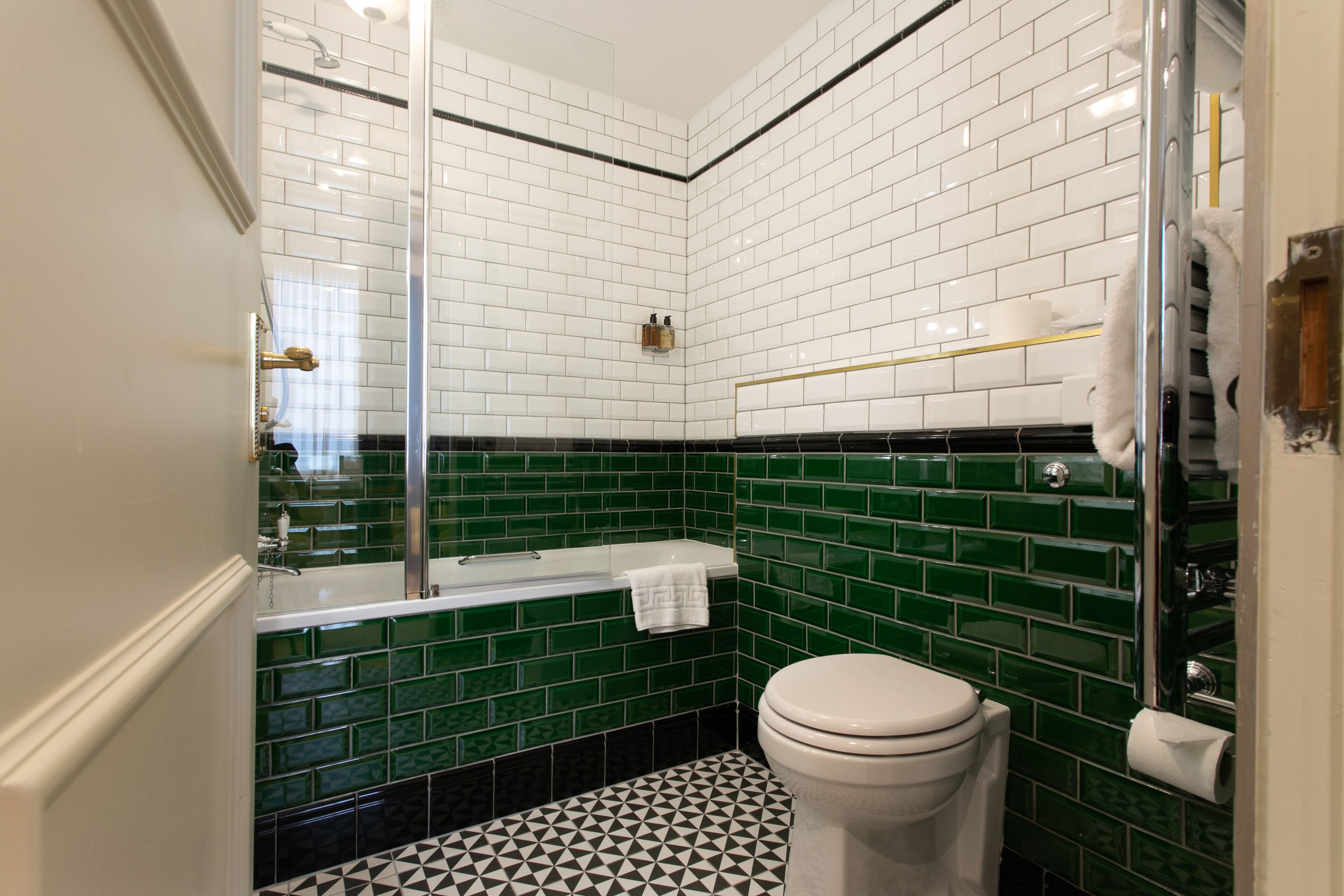 A bathroom with white and green tiled walls, a black and white patterned floor, a white toilet, and a glass-enclosed shower area.