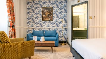 Habitación de hotel con un sofá azul, un sillón amarillo, una mesa de centro de madera y una cama blanca.