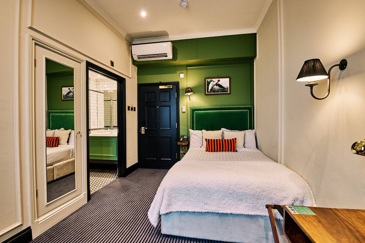 Habitación de hotel con cama, espejo, puerta que da a un baño y una lámpara fijada a la pared.