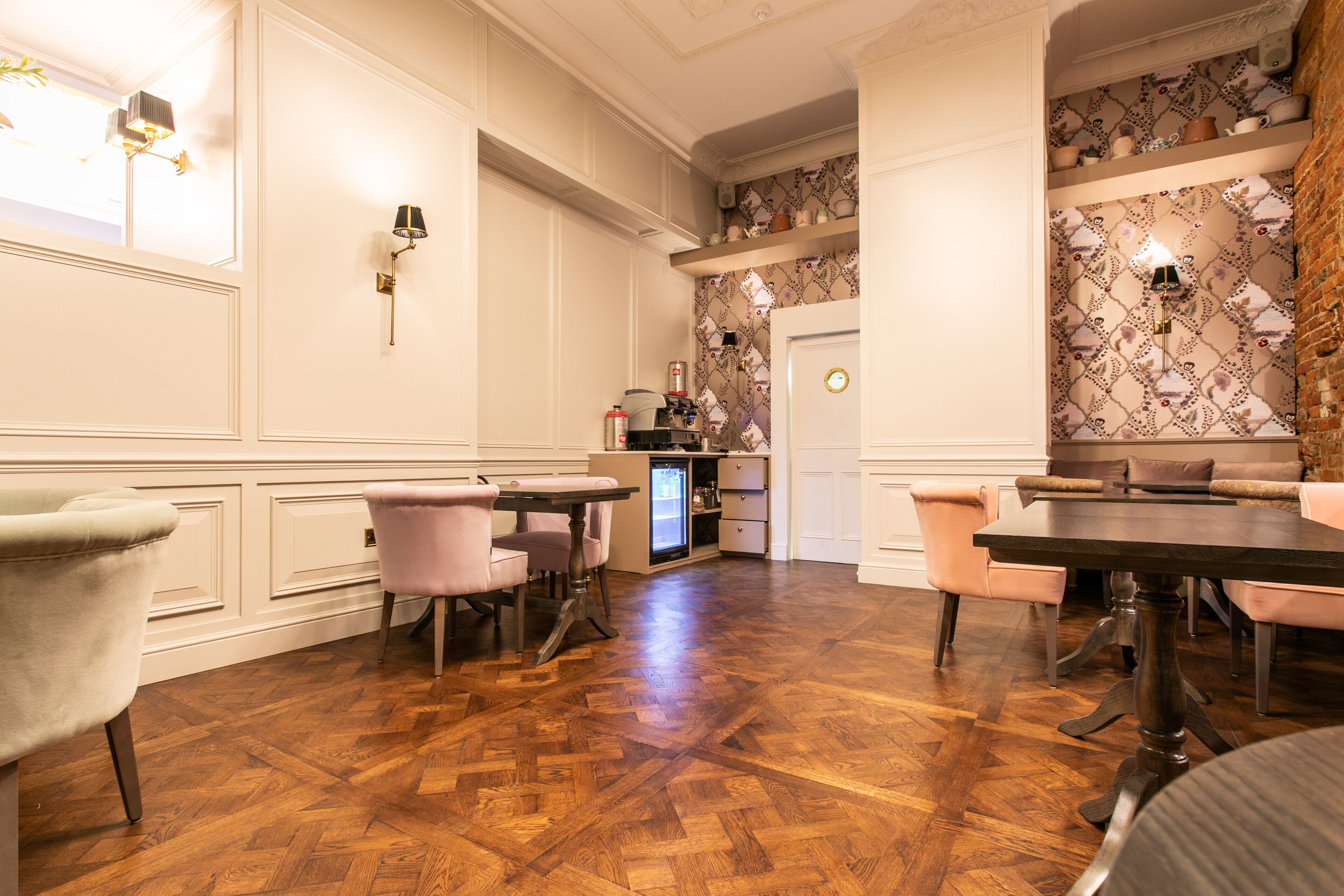 A dining area with a wooden herringbone floor, white paneled walls, and a patterned wallpaper on the back wall. There are two dining tables with chairs, and a refrigerator is visible in the background.