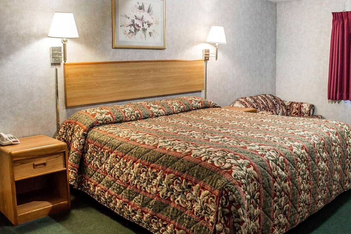 A hotel room with a wooden headboard, a floral-patterned bedspread, two bedside lamps, a nightstand with a phone, and a framed artwork on the wall.