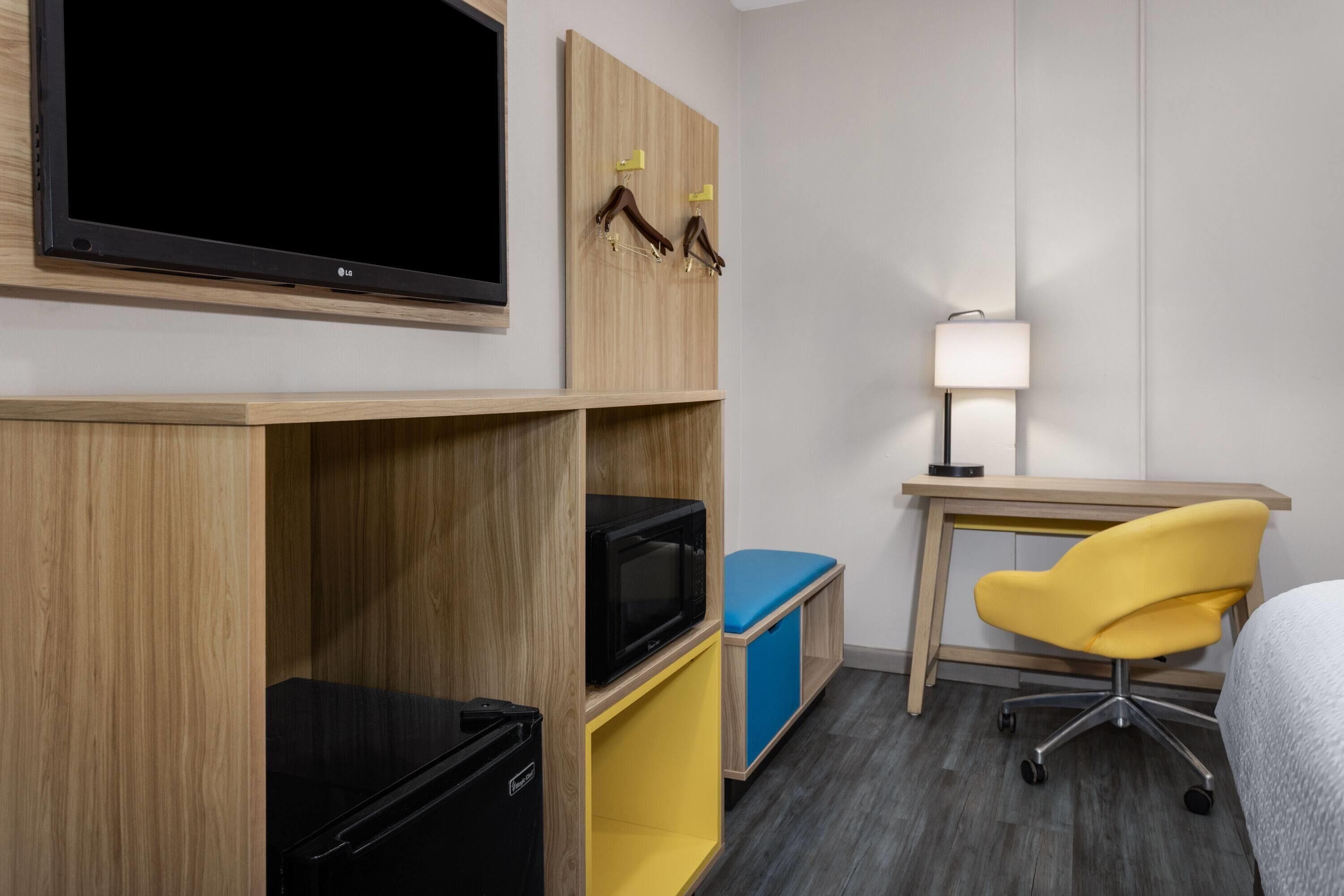 A hotel room with a flat-screen TV, wooden shelving, a desk with a lamp, a yellow chair, and a bed.