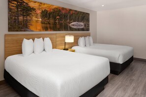 A hotel room with two beds, a wooden headboard, a painting of a lake and forest, and a nightstand with a lamp.