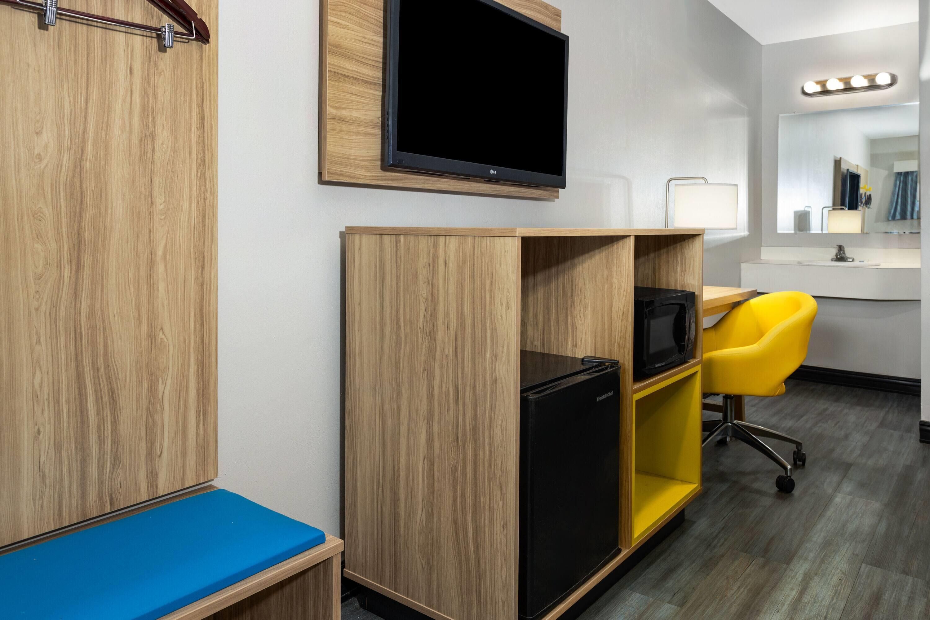 Een hotelkamer met houten meubels, een televisie, een kleine koelkast, een gele stoel en een spiegel met verlichting.