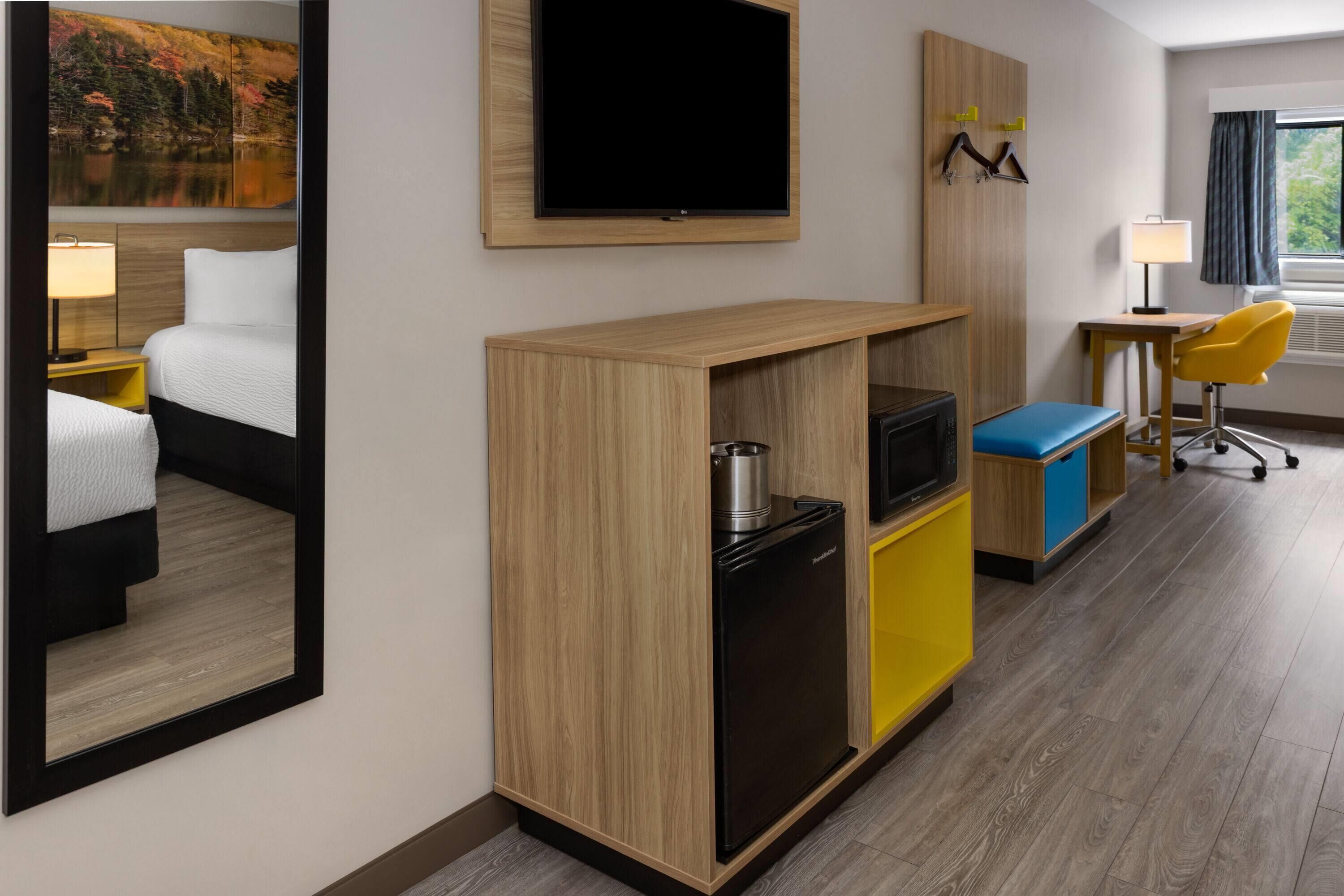 A hotel room with a bed, a TV, a small fridge, a desk with a chair, and a window with curtains.