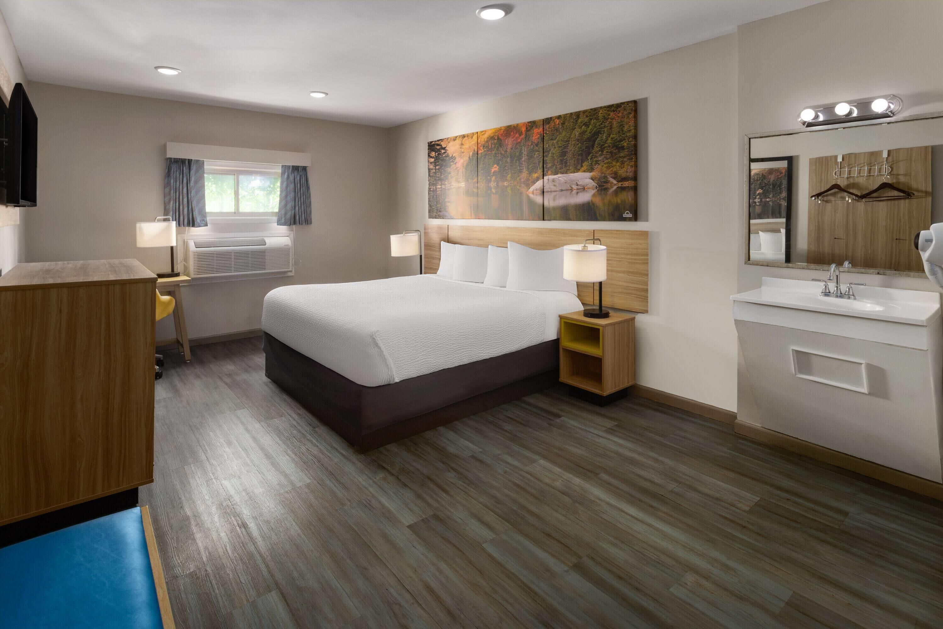 A hotel room with a bed, bedside lamps, a large window with curtains, a painting, a vanity with a mirror, and a wooden desk.