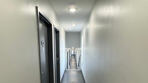 A long, narrow hallway with a dark wooden floor and white walls. There are doors on both sides of the hallway, and a vent is visible on the left wall.