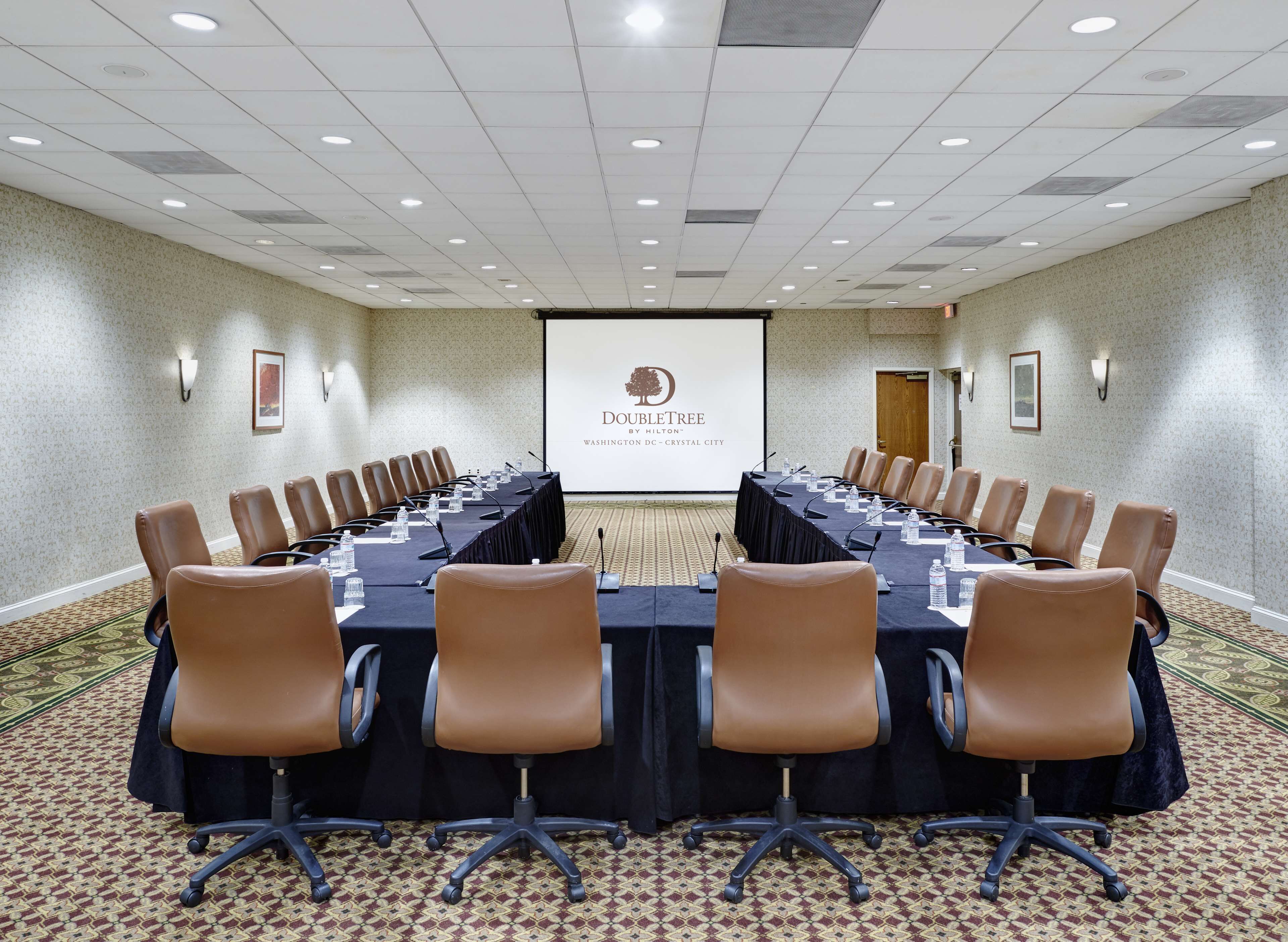 A conference room with a long table, brown leather chairs, and a projector screen displaying "DOUBLETREE by Hilton - Washington DC - Crystal City".
