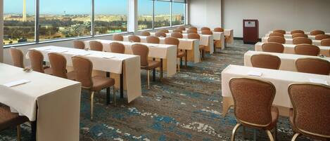 A conference room with tables and chairs, large windows with a city view, and a carpeted floor.