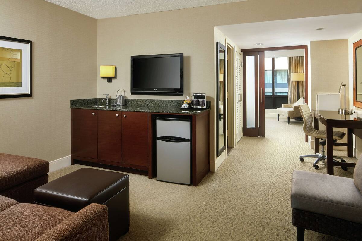 A hotel room with a kitchenette, a flat-screen TV, a desk with a chair, and a sofa.
