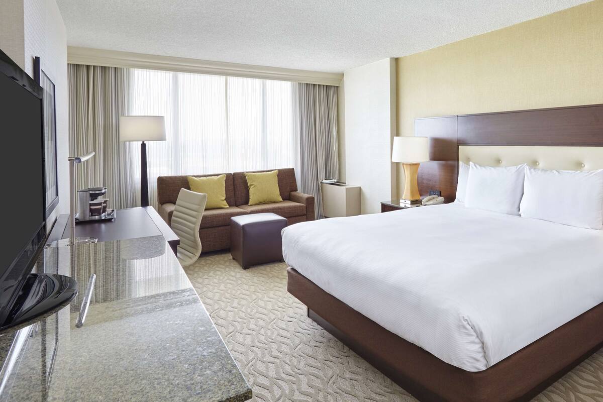 A hotel room with a large bed, a desk with a flat-screen TV, a sofa, and a nightstand with lamps.
