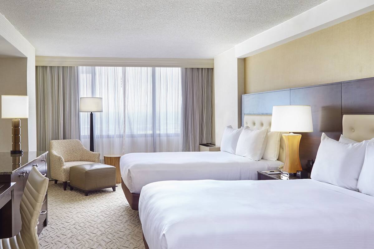 A hotel room with two beds, a desk, a chair, and a large window with curtains.