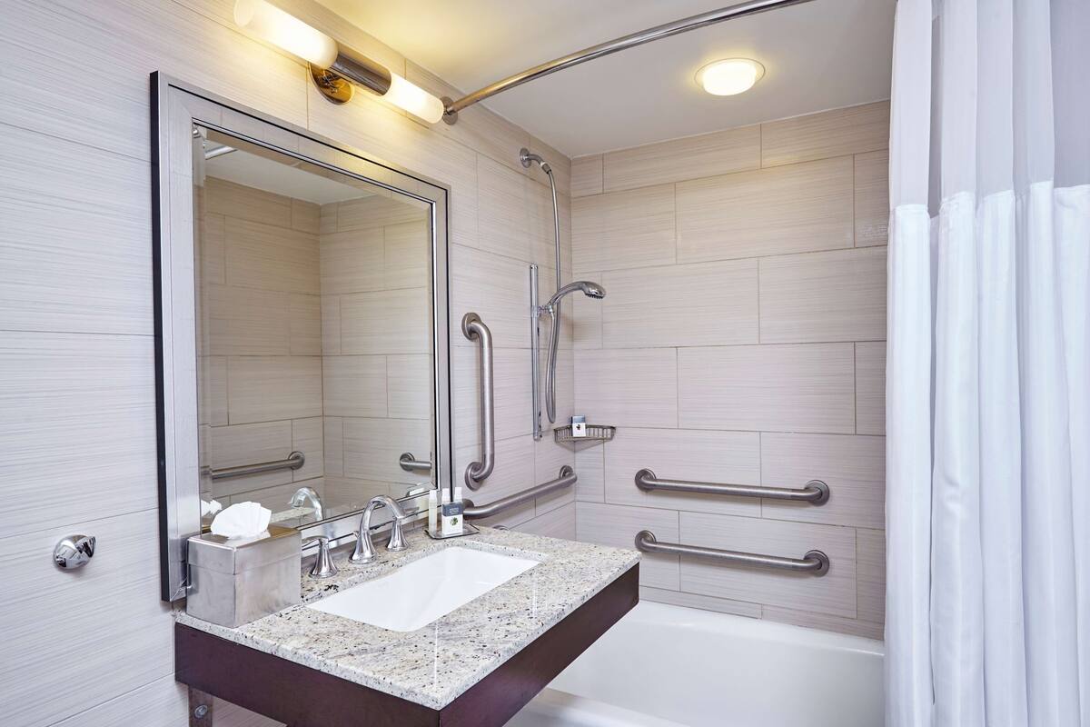A modern bathroom with a large mirror, a sink with a marble countertop, a bathtub, and handrails for accessibility.