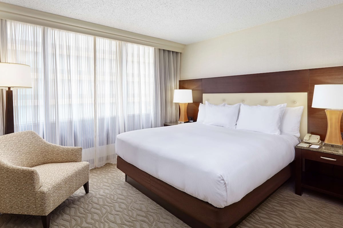A hotel room with a large bed, two bedside lamps, a chair, and a large window with sheer curtains.