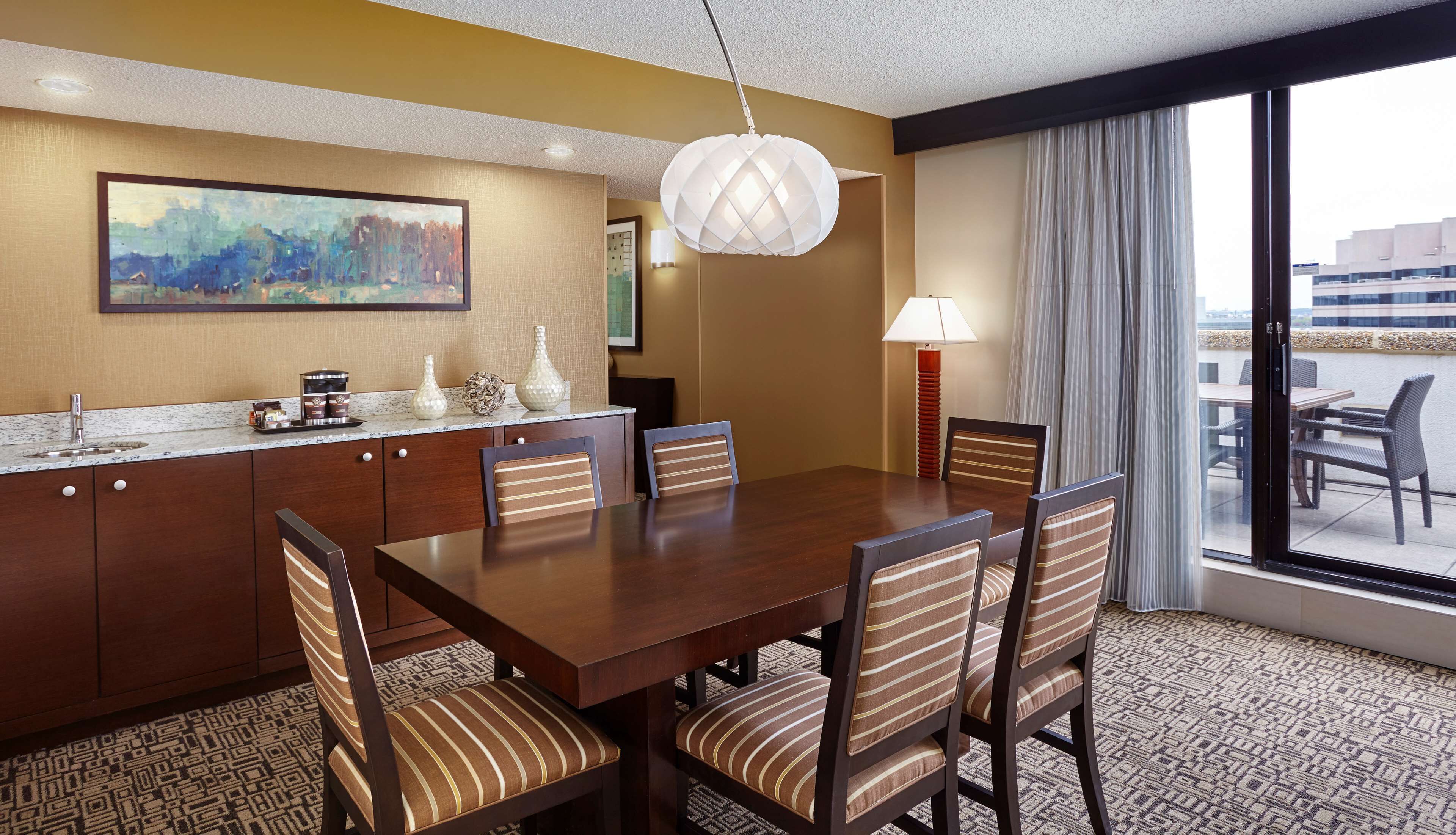A hotel dining area with a wooden table, striped chairs, a painting on the wall, a modern pendant light, and a view through large windows.