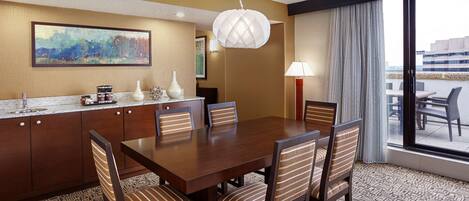 A hotel dining area with a wooden table, striped chairs, a painting on the wall, a modern pendant light, and a view through large windows.